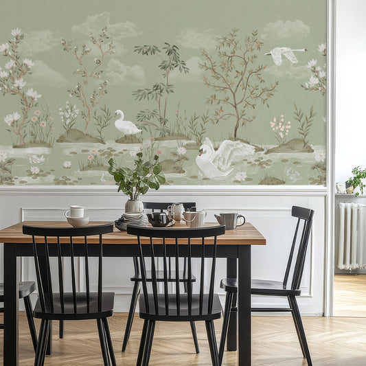 A dining room scene with a wooden table and black chairs in front of a large wall mural featuring a soft sage green landscape with swans, trees, and white clouds.
