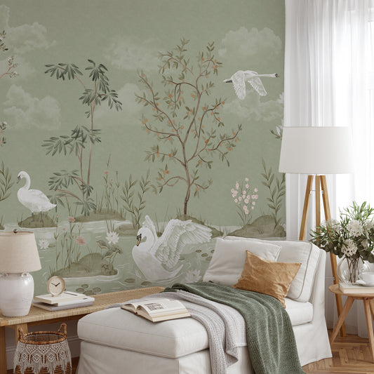 A styled living room featuring a sage green wallpaper with a delicate landscape design of swans, trees, and clouds. The room is furnished with a white armchair, a matching ottoman, a white floor lamp, and a side table with flowers.