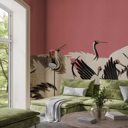 A styled living room featuring a prominent pink wall with a large-scale crane and bird pattern wallpaper. In front of the wall is a green sectional sofa with matching green cushions, a white throw blanket, a floor lamp, and a round wooden coffee table with books and a glass vase.