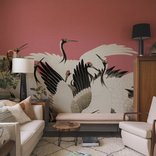 A styled living room featuring a large wall mural of white cranes with black and red details on a dusty rose pink wall. The room includes a cream sofa, a wooden coffee table, a white armchair, and a wooden cabinet.