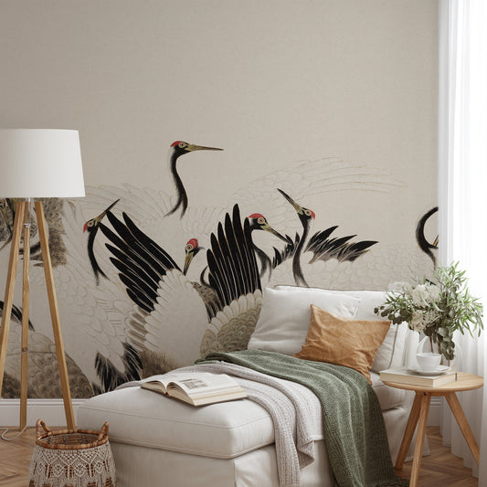 A styled bedroom scene featuring a large wall mural of black and white cranes with red accents on a beige wall. The room includes a cream-colored ottoman with a green throw blanket, a wooden floor lamp, a small wooden side table with a vase of flowers, and white pillows.