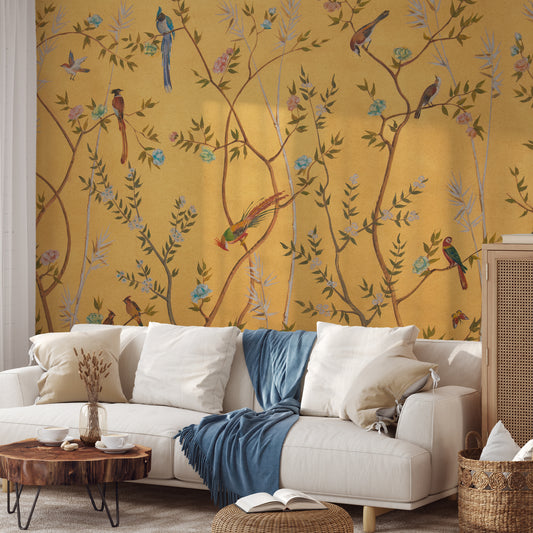 A styled living room featuring a bold mustard yellow wallpaper with a detailed pattern of birds perched on branches with leaves. In front of the wall is a white sofa with white and beige pillows, a blue throw blanket, a rustic wooden coffee table, and a woven rattan cabinet.