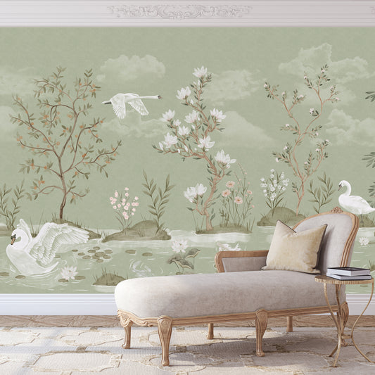 A styled interior scene showing a pale sage green wall mural with a delicate botanical and watercolor landscape, including swans, flowering trees, clouds, and a chaise lounge in front of it.